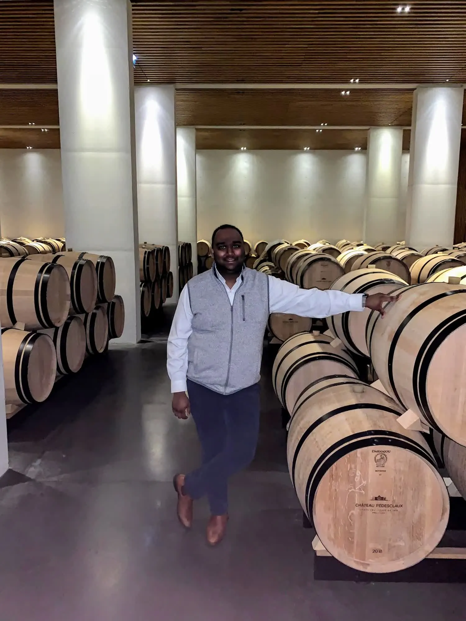 Yagna Vudathu standing among oak barrels in a Pauillac barrel hall
